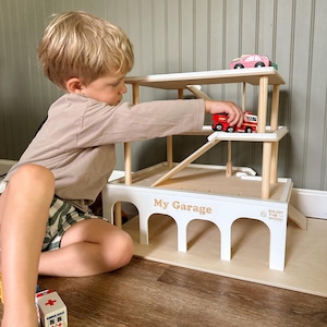 K&ouml;nnte beinhalten: Ein wei&szlig;es Holzspielzeug-Parkhaus mit der Aufschrift "My Garage". Es hat nat&uuml;rliche Holzakzente, eine Rampe und mehrere Ebenen. Ein Kind spielt mit dem Spielzeug und platziert einen roten Feuerwehrauto auf der Rampe.