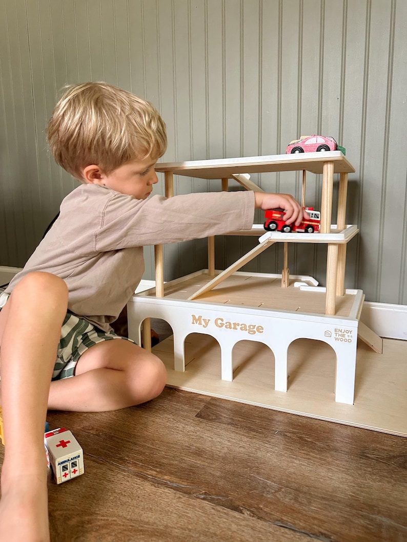 K&ouml;nnte beinhalten: Ein wei&szlig;es Holzspielzeug-Parkhaus mit der Aufschrift "My Garage". Es hat nat&uuml;rliche Holzakzente, eine Rampe und mehrere Ebenen. Ein Kind spielt mit dem Spielzeug und platziert einen roten Feuerwehrauto auf der Rampe.