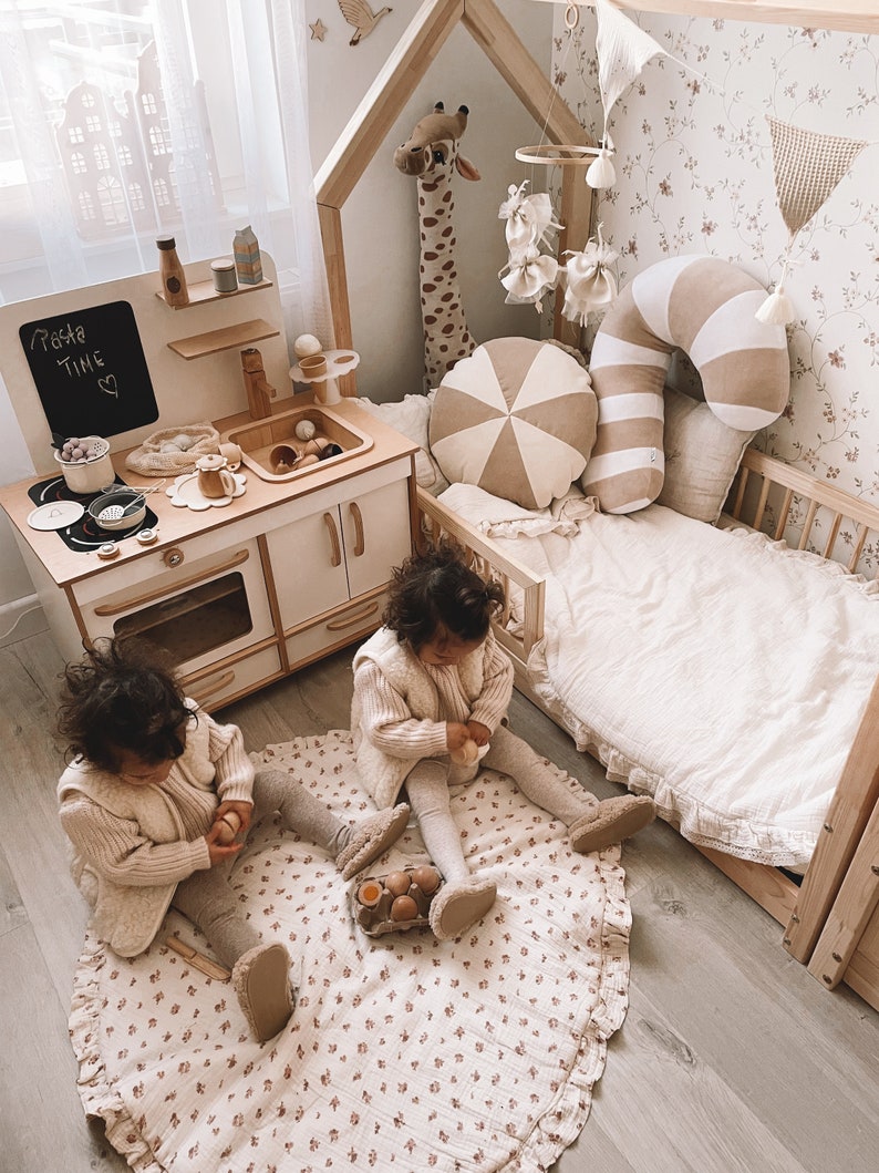 May include: Two children play with wooden eggs on a floral play rug in a room with a play kitchen and a wooden bed. The play kitchen has a chalkboard sign that says "Pasta Time".