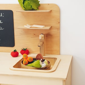 May include: A wooden play kitchen with a sink, two shelves, and a chalkboard. The shelves are filled with toy vegetables and a plastic fork and knife. The sink is made of white plastic and has a wooden faucet.