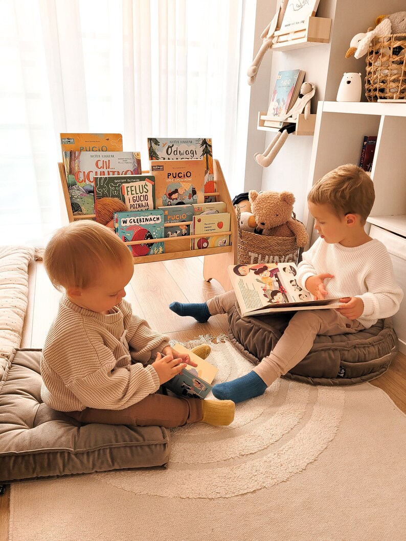 May include: Two children sit on plush cushions reading books. The child on the left is holding a book with a blue cover. The child on the right is holding a book with a colorful cover. Both children are wearing brown pants and socks. The bookshelves behind them are filled with colorful books. The bookshelves are made of wood and have a light brown finish.