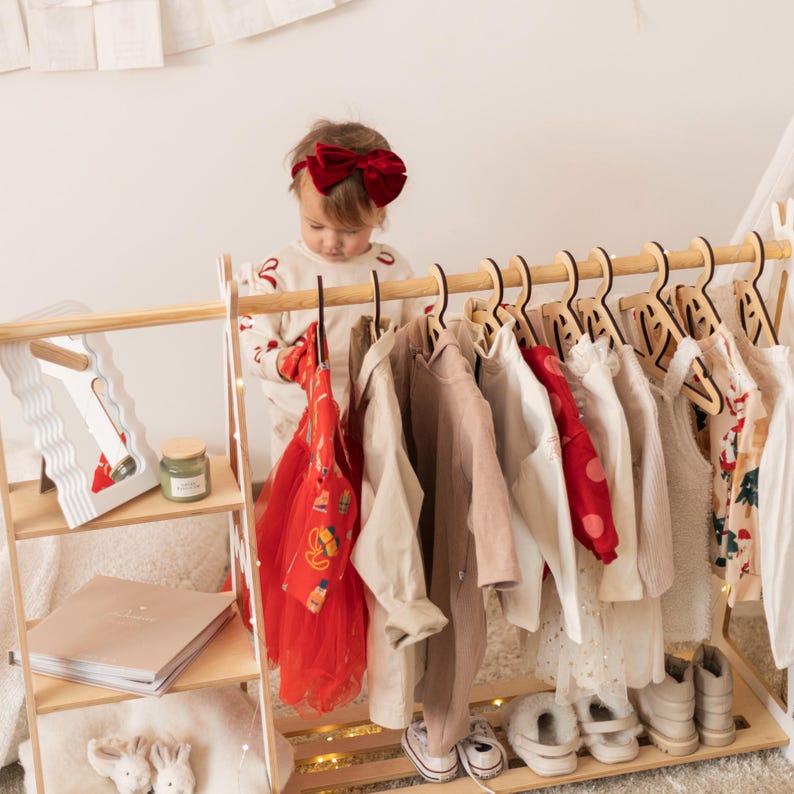 May include: A wooden children's clothing rack with a variety of clothes hanging on it. A young child is standing in front of the rack, looking at the clothes. The rack has a shelf on the bottom and a shelf on the top. The rack is painted white.