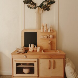 May include: A wooden play kitchen with a black chalkboard, a sink, an oven, and two cabinets. The kitchen is made of light wood and has a white countertop. There are several toy food items and utensils on the countertop. A white pillow is on the floor next to the kitchen.
