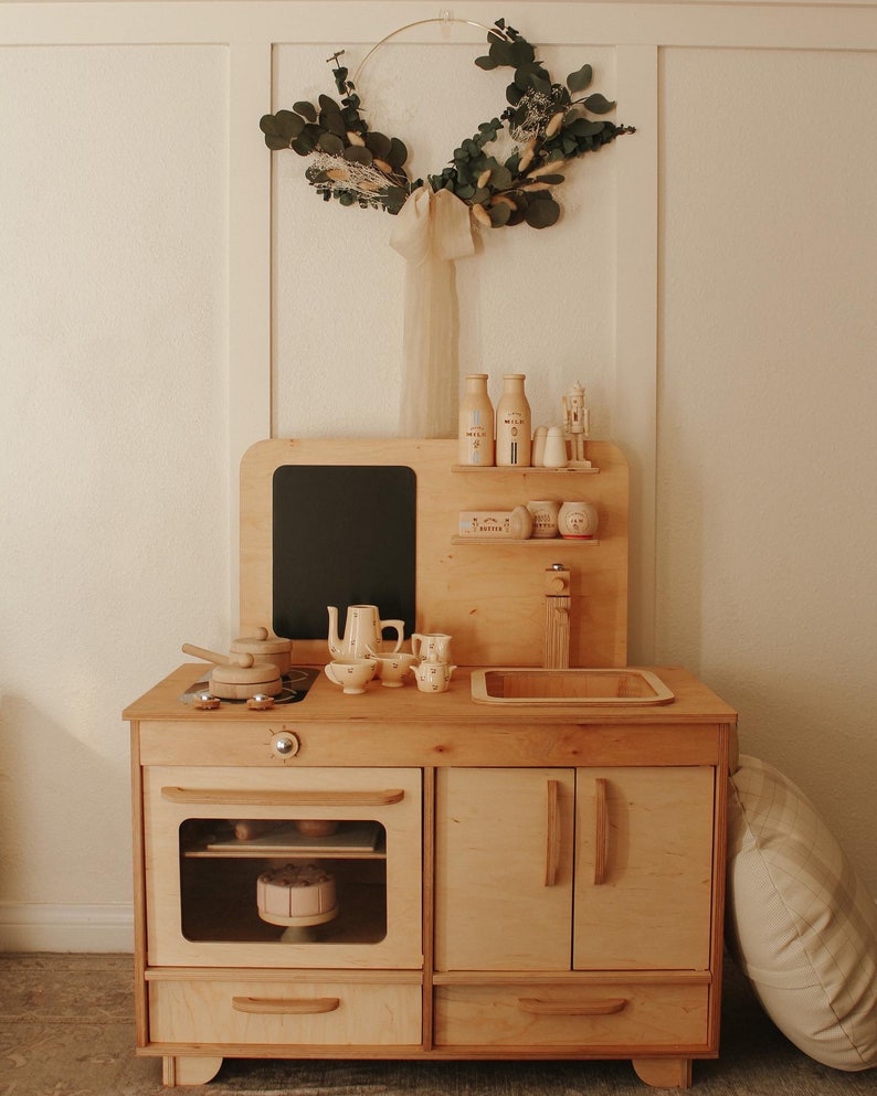 May include: A wooden play kitchen with a black chalkboard, a sink, an oven, and two cabinets. The kitchen is made of light wood and has a white countertop. There are several toy food items and utensils on the countertop. A white pillow is on the floor next to the kitchen.