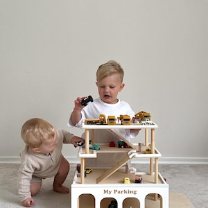 K&ouml;nnte beinhalten: Ein mehrst&ouml;ckiges Spielzeug-Parkhaus aus Holz mit Rampen und Bogeneing&auml;ngen. Das Parkhaus ist wei&szlig; mit nat&uuml;rlichen Holzdetails und dem Text "My Parking". Kleine Spielzeugfahrzeuge sind auf den Ebenen angeordnet. Zwei Kinder spielen mit dem Spielzeug.