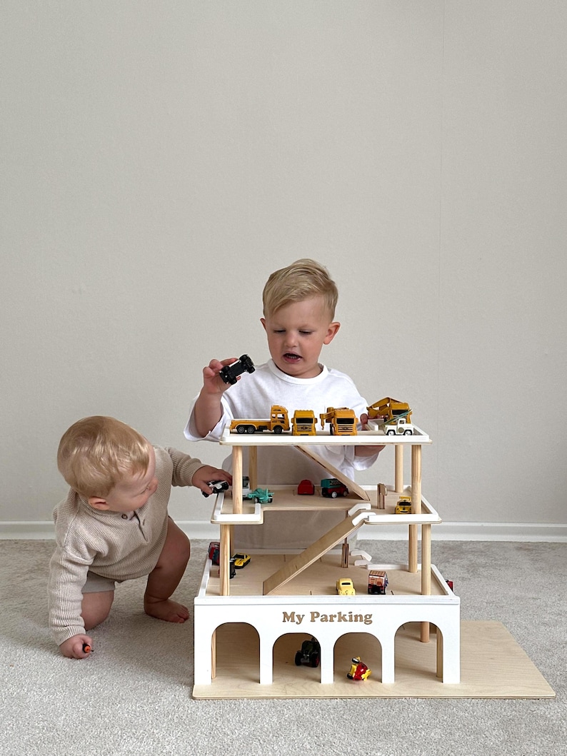 K&ouml;nnte beinhalten: Ein mehrst&ouml;ckiges Spielzeug-Parkhaus aus Holz mit Rampen und Bogeneing&auml;ngen. Das Parkhaus ist wei&szlig; mit nat&uuml;rlichen Holzdetails und dem Text "My Parking". Kleine Spielzeugfahrzeuge sind auf den Ebenen angeordnet. Zwei Kinder spielen mit dem Spielzeug.