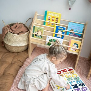 May include: A light-colored wooden bookshelf holding five children's books with colorful covers, positioned next to a woven basket and a white wooden alphabet puzzle board featuring multicolored letters and the word Alphabet.