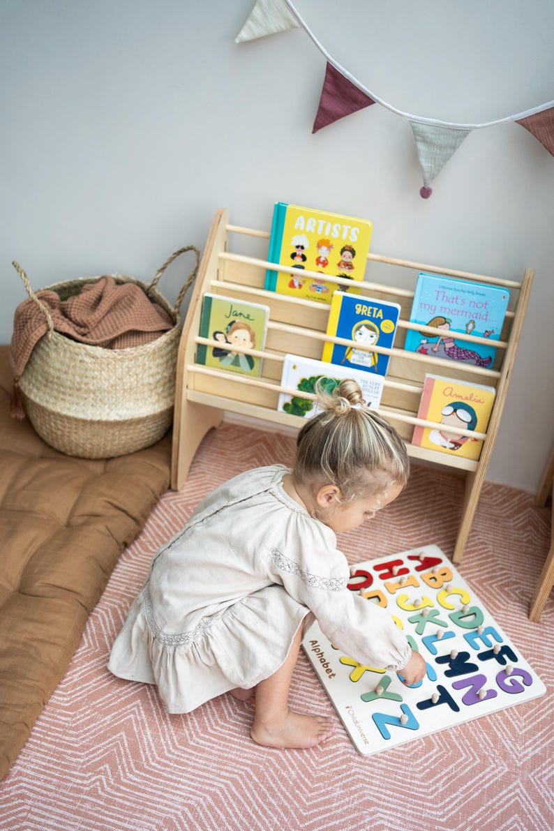 May include: A light-colored wooden bookshelf holding five children's books with colorful covers, positioned next to a woven basket and a white wooden alphabet puzzle board featuring multicolored letters and the word Alphabet.