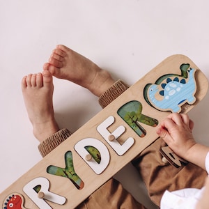 May include: Wooden name puzzle spelling "RYDER" with white letters and dinosaur illustrations. The puzzle has a light wood finish and includes a red and blue dinosaur. A child's hands and feet are visible.