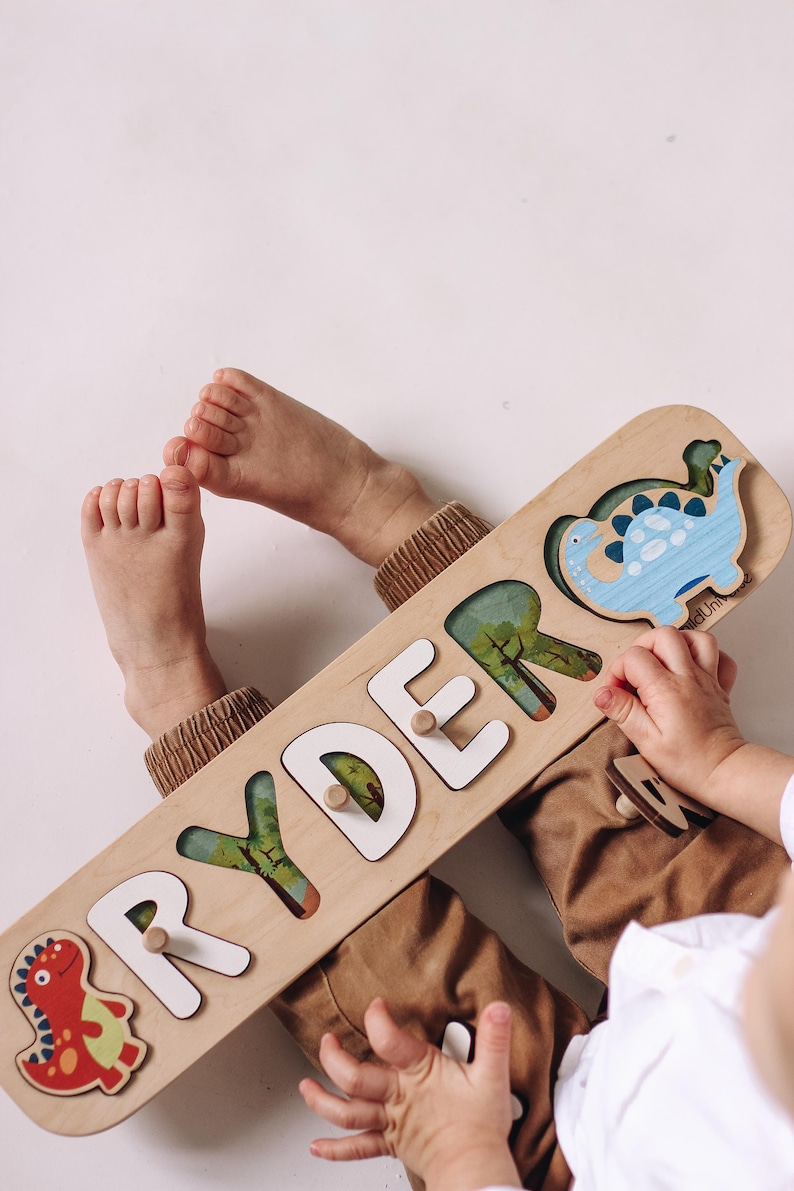 May include: Wooden name puzzle spelling "RYDER" with white letters and dinosaur illustrations. The puzzle has a light wood finish and includes a red and blue dinosaur. A child's hands and feet are visible.