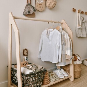 May include: A wooden clothing rack with a shelf and a basket. The rack is made of light wood and has a simple design. There are clothes hanging on the rack, including a white shirt and a plaid shirt. There is a basket on the shelf with folded clothes and a basket on the floor with a stuffed animal.