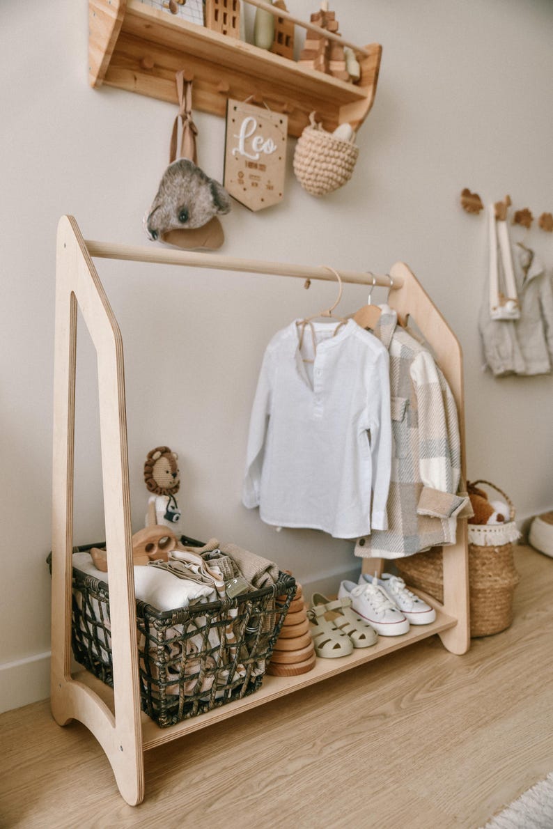 May include: A wooden clothing rack with a shelf and a basket. The rack is made of light wood and has a simple design. There are clothes hanging on the rack, including a white shirt and a plaid shirt. There is a basket on the shelf with folded clothes and a basket on the floor with a stuffed animal.