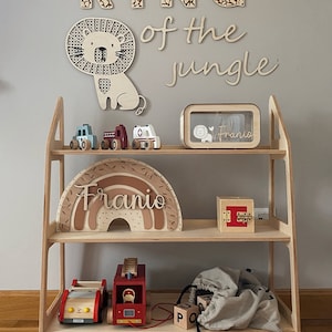 May include: A wooden shelf unit with two shelves, featuring a wooden lion cutout, wooden letters spelling "KING of the jungle", a wooden rainbow with the name "Franio" on it, a wooden toy fire truck, a wooden toy car, a wooden toy ambulance, a wooden box with a lock, and a wooden box with a glass front and the name "Franio" on it.