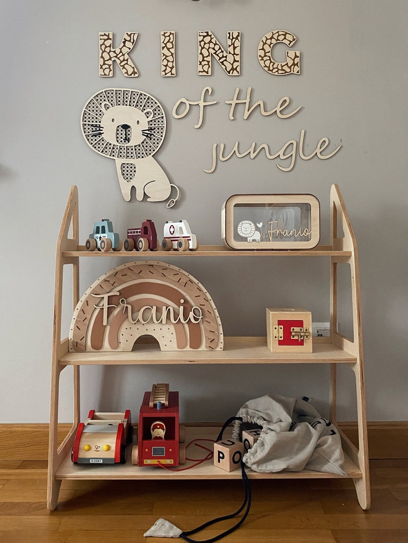 May include: A wooden shelf unit with two shelves, featuring a wooden lion cutout, wooden letters spelling "KING of the jungle", a wooden rainbow with the name "Franio" on it, a wooden toy fire truck, a wooden toy car, a wooden toy ambulance, a wooden box with a lock, and a wooden box with a glass front and the name "Franio" on it.