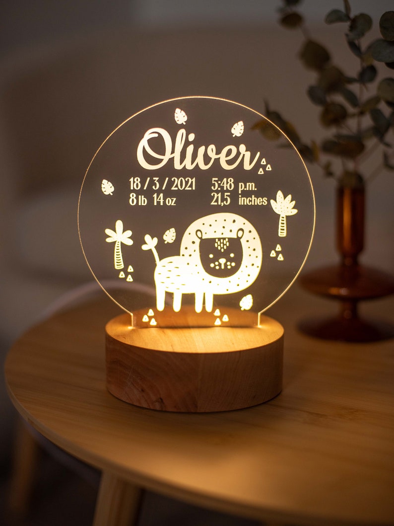 May include: A personalised night light with a lion design. The clear acrylic light features the name "Oliver", birthdate, time, weight (3.9 kg), and height (54.6 cm). The light sits on a wooden base and emits a warm glow.