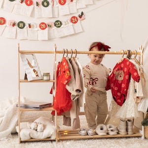 Puede incluir: Un perchero de madera con un estante blanco y una alfombra blanca y esponjosa debajo. Un ni&ntilde;o peque&ntilde;o est&aacute; de pie frente al perchero, con una camisa de manga larga de color crema con un lazo rojo y pantalones beige. El perchero est&aacute; lleno de varias prendas de vestir, incluyendo un vestido rojo, un vestido blanco y una camisa con estampado rojo y blanco.
