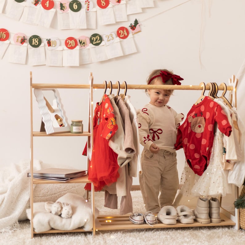 Puede incluir: Un perchero de madera con un estante blanco y una alfombra blanca y esponjosa debajo. Un ni&ntilde;o peque&ntilde;o est&aacute; de pie frente al perchero, con una camisa de manga larga de color crema con un lazo rojo y pantalones beige. El perchero est&aacute; lleno de varias prendas de vestir, incluyendo un vestido rojo, un vestido blanco y una camisa con estampado rojo y blanco.
