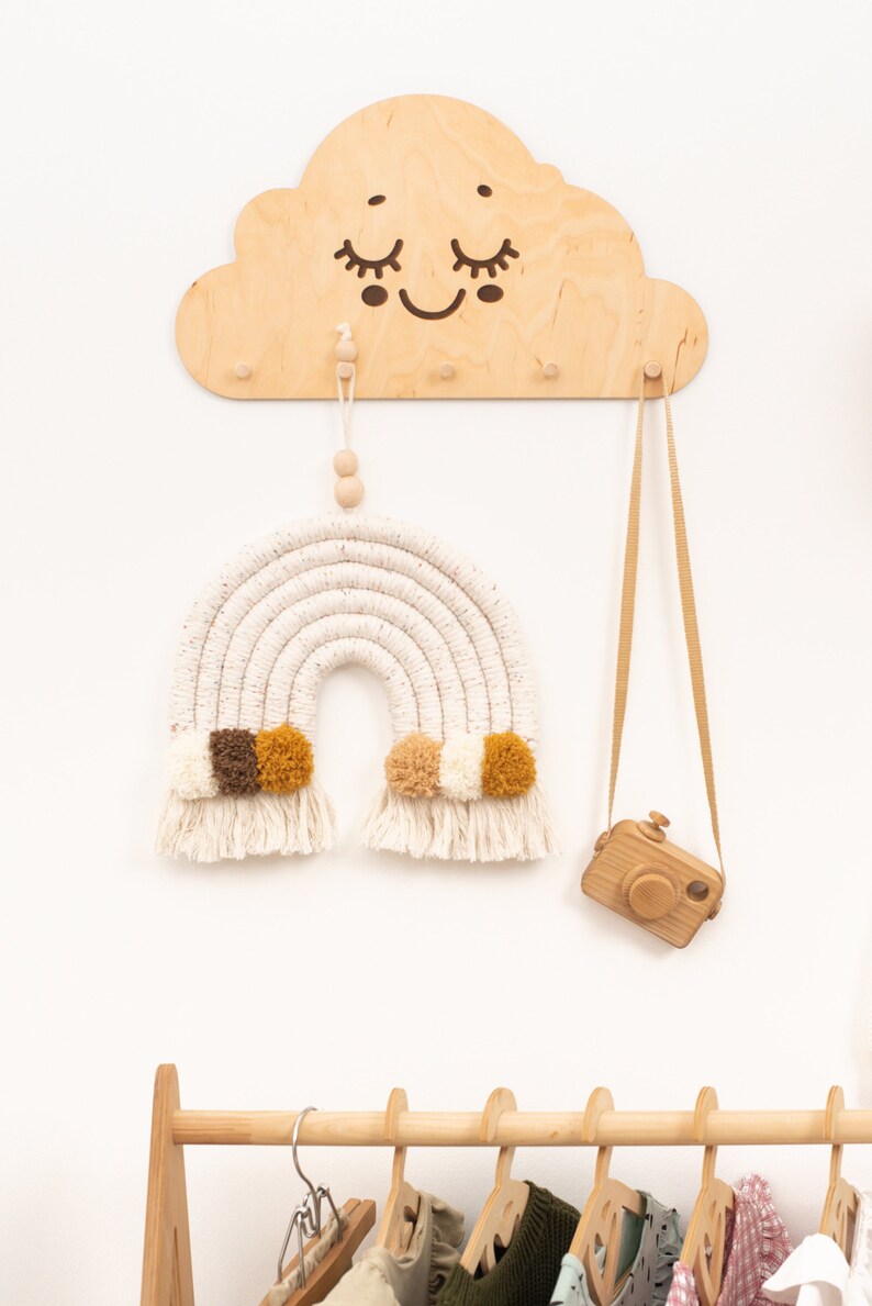 May include: A wooden cloud-shaped wall hook with a smiling face and three hooks. A macrame rainbow with brown and yellow pom-poms hangs from one hook. A wooden camera with a brown strap hangs from another hook. A wooden clothes rack with several hangers and clothes is in the background.