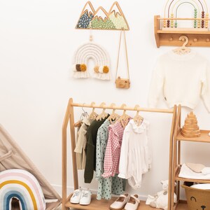 May include: A wooden clothing rack with a white, pink, and green patterned shirt, a green sweater, and a white dress hanging on it. There are also two pairs of white shoes on the bottom shelf. The rack is in a room with a white wall and a wooden shelf with a rainbow abacus on it.