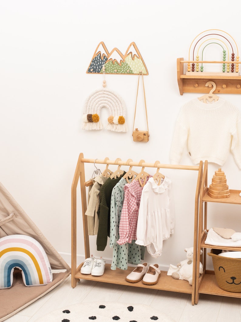 May include: A wooden clothing rack with a white, pink, and green patterned shirt, a green sweater, and a white dress hanging on it. There are also two pairs of white shoes on the bottom shelf. The rack is in a room with a white wall and a wooden shelf with a rainbow abacus on it.