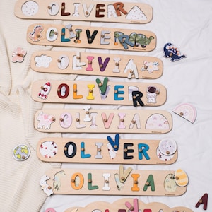 May include: Wooden name puzzles spelling "OLIVER" and "OLIVIA" in various colors. Each puzzle has a unique design, with decorative elements like dinosaurs, rockets, and rainbows. The puzzles are on a white surface.