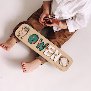 May include: Wooden name puzzle spelling "OWEN" with baseball and astronaut illustrations. The letters are white, and the "O" is teal with a baseball design. The puzzle is made by ChildUniverse.