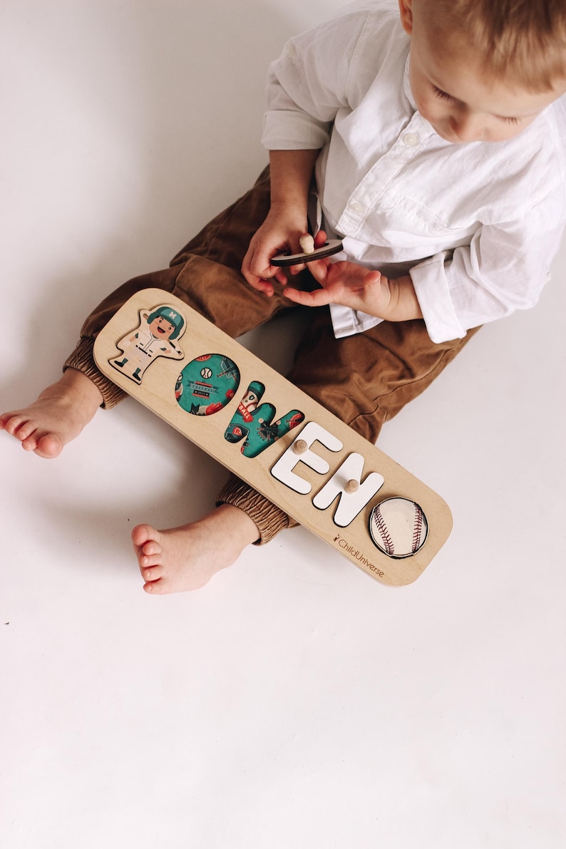 May include: Wooden name puzzle spelling "OWEN" with baseball and astronaut illustrations. The letters are white, and the "O" is teal with a baseball design. The puzzle is made by ChildUniverse.