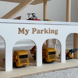 K&ouml;nnte beinhalten: Ein mehrst&ouml;ckiges Spielzeug-Parkhaus aus Holz mit Rampen. Die wei&szlig;e Fassade tr&auml;gt den Text "My Parking" in braunen Buchstaben. Mehrere Spielzeug-Lkw und Baufahrzeuge sind in der Garage geparkt.