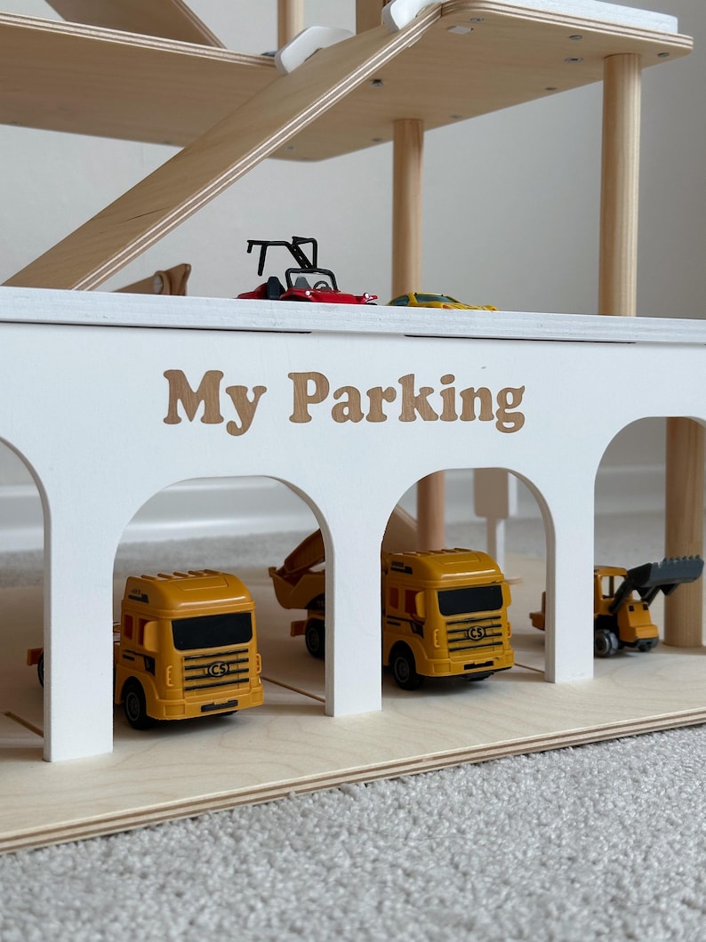 K&ouml;nnte beinhalten: Ein mehrst&ouml;ckiges Spielzeug-Parkhaus aus Holz mit Rampen. Die wei&szlig;e Fassade tr&auml;gt den Text "My Parking" in braunen Buchstaben. Mehrere Spielzeug-Lkw und Baufahrzeuge sind in der Garage geparkt.