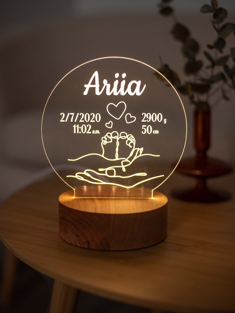 May include: A personalised night light with the name "Ariia" illuminated in white. The acrylic light displays birth details: date, time, weight (2900g), and length (50cm). The design includes hearts and baby feet, set on a wooden base.