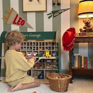 K&ouml;nnte beinhalten: Ein Kind spielt mit Spielzeugautos in einer Garage-Themen-Vitrine aus Holz. Die Vitrine ist gr&uuml;n und hat mehrere F&auml;cher, die mit bunten Spielzeugfahrzeugen gef&uuml;llt sind. Ein roter Cowboyhut h&auml;ngt in der N&auml;he, und ein Teddyb&auml;r sitzt auf einem Tisch.