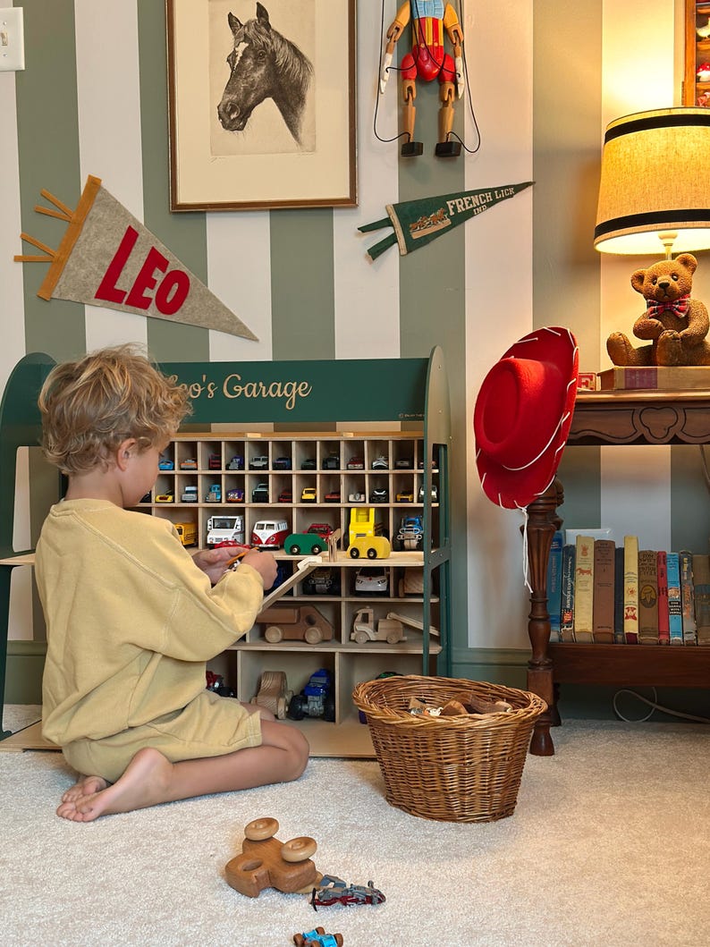 K&ouml;nnte beinhalten: Ein Kind spielt mit Spielzeugautos in einer Garage-Themen-Vitrine aus Holz. Die Vitrine ist gr&uuml;n und hat mehrere F&auml;cher, die mit bunten Spielzeugfahrzeugen gef&uuml;llt sind. Ein roter Cowboyhut h&auml;ngt in der N&auml;he, und ein Teddyb&auml;r sitzt auf einem Tisch.