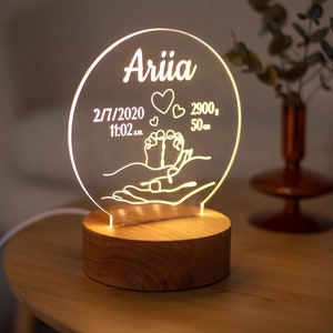 May include: A personalised night light featuring the name "Ariia" on a clear, circular acrylic panel. The panel displays the birth date, time, weight (2.9 kg), and length (50 cm) of a baby, along with heart and foot symbols. It rests on a round wooden base.
