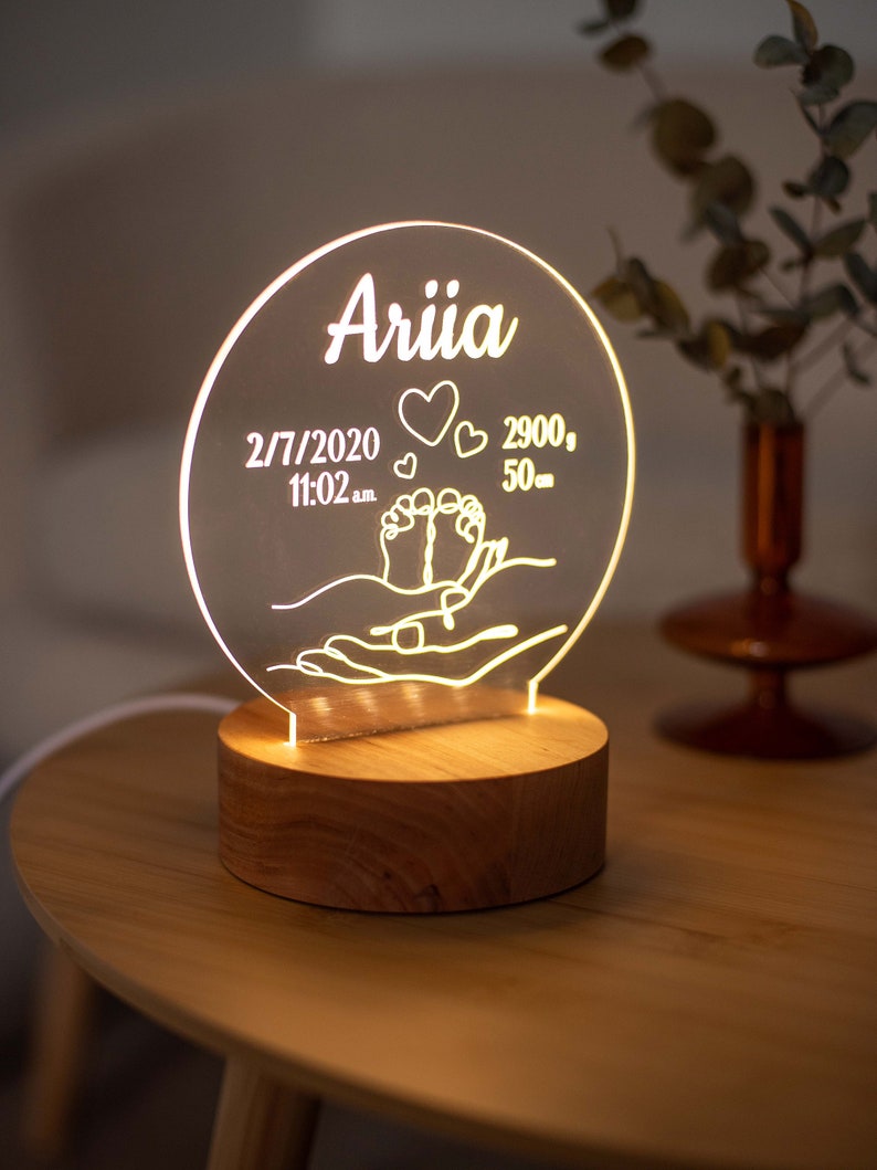May include: A personalised night light featuring the name "Ariia" on a clear, circular acrylic panel. The panel displays the birth date, time, weight (2.9 kg), and length (50 cm) of a baby, along with heart and foot symbols. It rests on a round wooden base.