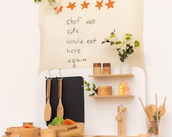 May include: A wooden toy kitchen set with a white countertop and a chalkboard. The set includes wooden utensils, a sink, and a backdrop with the text "chef was cute. would eat here again."