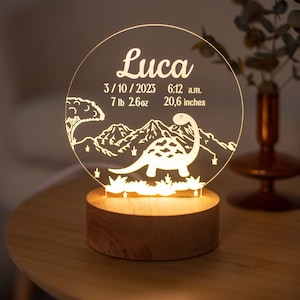 May include: A personalised night light featuring the name "Luca" on a clear acrylic disc. The design includes a dinosaur, mountains, and the date 3/10/2023, time 6:12 a.m., weight 3.2 kg, and height 52.3 cm. The light sits on a wooden base.