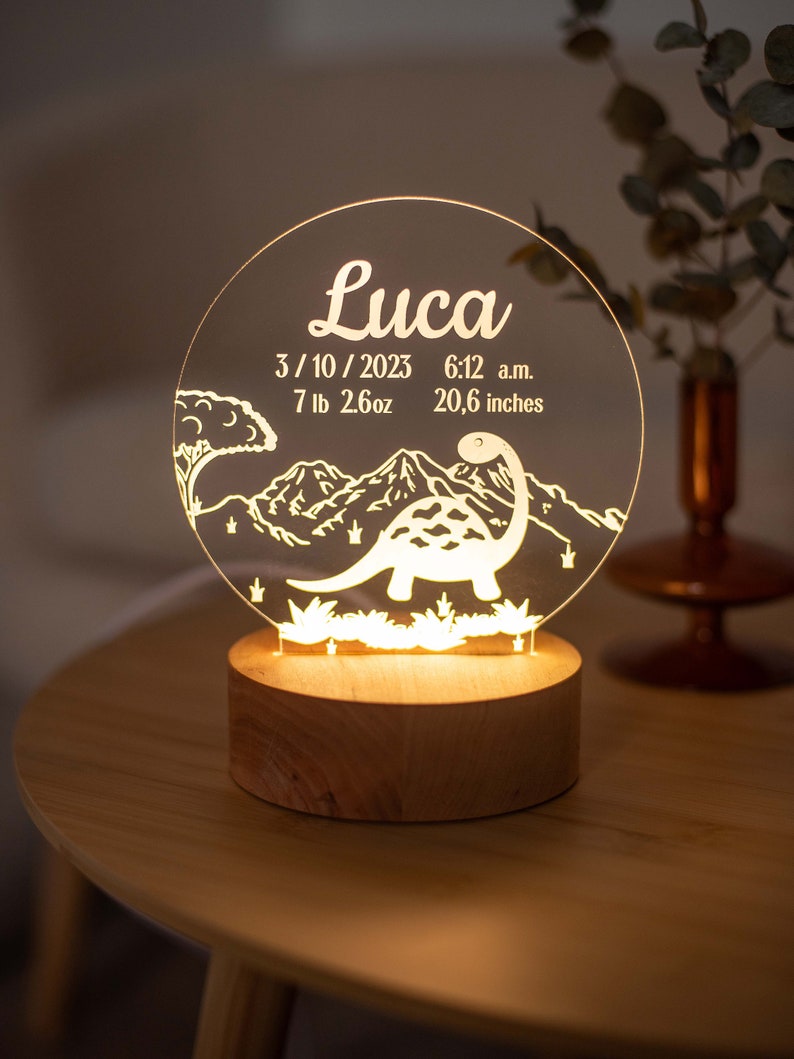 May include: A personalised night light featuring the name "Luca" on a clear acrylic disc. The design includes a dinosaur, mountains, and the date 3/10/2023, time 6:12 a.m., weight 3.2 kg, and height 52.3 cm. The light sits on a wooden base.