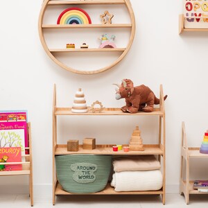 May include: A wooden shelf unit with three shelves, a green storage basket labeled "Around the World", and a brown stuffed dinosaur. The shelf unit is against a white wall with a round wooden shelf with rainbow and star-shaped toys on it.