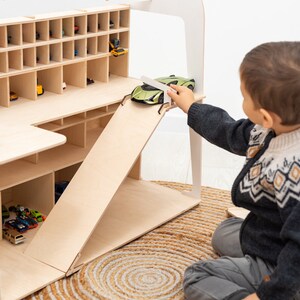 K&ouml;nnte beinhalten: Ein Holzspielzeug-Garage mit mehreren F&auml;chern f&uuml;r Spielzeugautos. Die Garage hat eine Rampe und den Text "Oliver's Garage" oben. Ein Kind spielt mit einem gr&uuml;nen Spielzeugauto auf der Rampe. Die Garage steht auf einem gewebten Teppich.