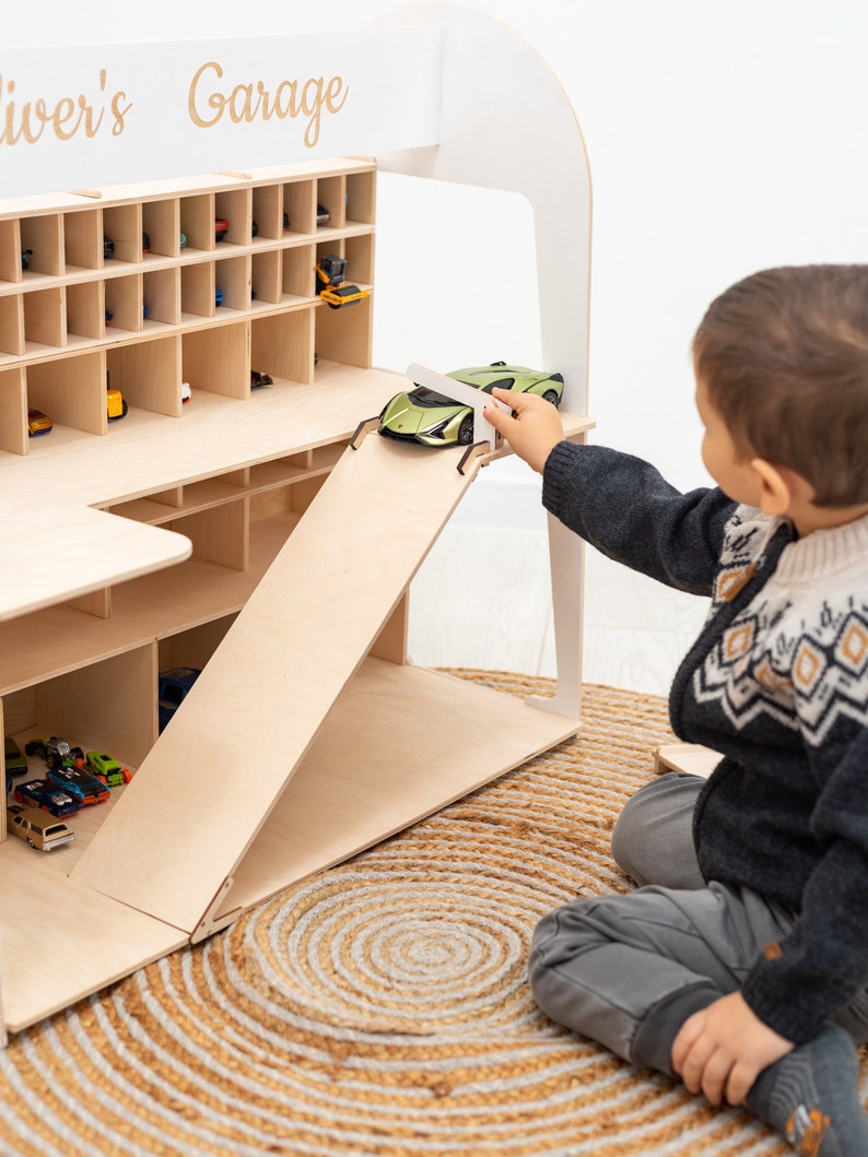 K&ouml;nnte beinhalten: Ein Holzspielzeug-Garage mit mehreren F&auml;chern f&uuml;r Spielzeugautos. Die Garage hat eine Rampe und den Text "Oliver's Garage" oben. Ein Kind spielt mit einem gr&uuml;nen Spielzeugauto auf der Rampe. Die Garage steht auf einem gewebten Teppich.