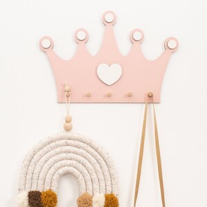 May include: A pink wooden crown with a white heart in the center, hanging on a white wall. There are three wooden hooks on the crown. A white and brown macrame rainbow with brown and yellow pom-poms is hanging from one hook. A wooden camera with a brown strap is hanging from another hook.
