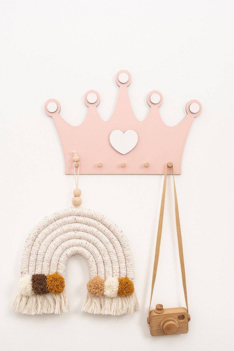 May include: A pink wooden crown with a white heart in the center, hanging on a white wall. There are three wooden hooks on the crown. A white and brown macrame rainbow with brown and yellow pom-poms is hanging from one hook. A wooden camera with a brown strap is hanging from another hook.