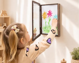 May include: A rectangular dark wood hinged display frame mounted on a wall, featuring a clear front panel that opens to reveal a white paper insert with a hand-painted illustration of three colorful flowers in yellow, pink, and light blue.