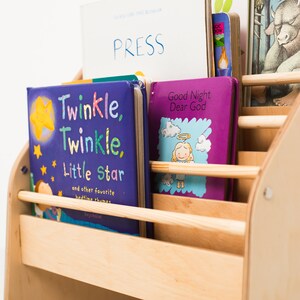 May include: A light-colored wooden bookshelf with horizontal slats holding several children's books. The front books are titled Twinkle, Twinkle, Little Star and Good Night Dear God. Behind them, a book with the word PRESS is visible.