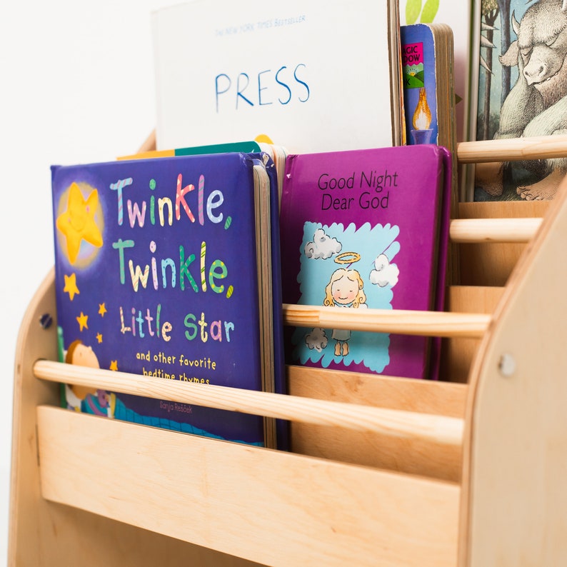 May include: A light-colored wooden bookshelf with horizontal slats holding several children's books. The front books are titled Twinkle, Twinkle, Little Star and Good Night Dear God. Behind them, a book with the word PRESS is visible.