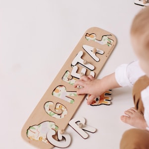 May include: Wooden puzzle spelling out the name "GRETA" with white letters and safari animal illustrations. The puzzle is on a light-colored surface, with a child's hand interacting with the puzzle.