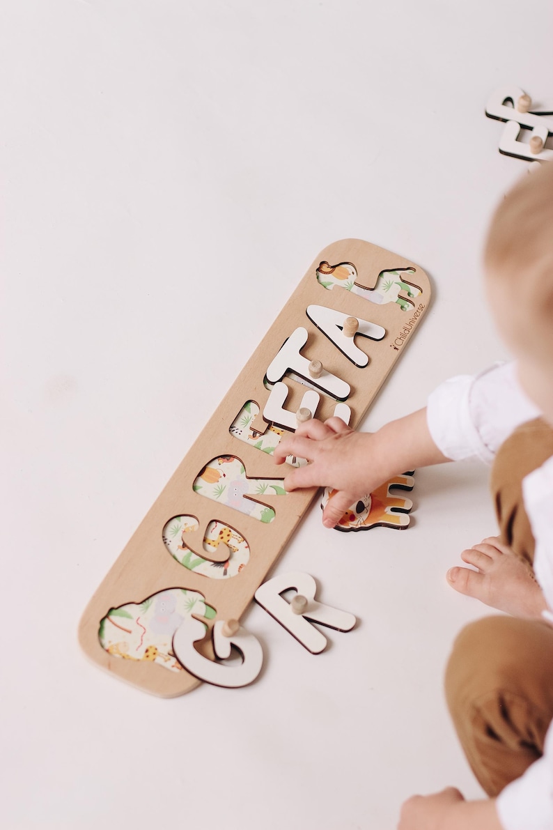 May include: Wooden puzzle spelling out the name "GRETA" with white letters and safari animal illustrations. The puzzle is on a light-colored surface, with a child's hand interacting with the puzzle.