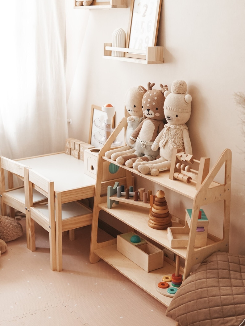 May include: A wooden shelf unit with three shelves, filled with toys and stuffed animals. The top shelf has three stuffed animals, a deer, a rabbit, and a bear. The middle shelf has a wooden toy train track, a wooden stacking toy, and a wooden abacus. The bottom shelf has a wooden box, a wooden ring stack, and a wooden toy car.