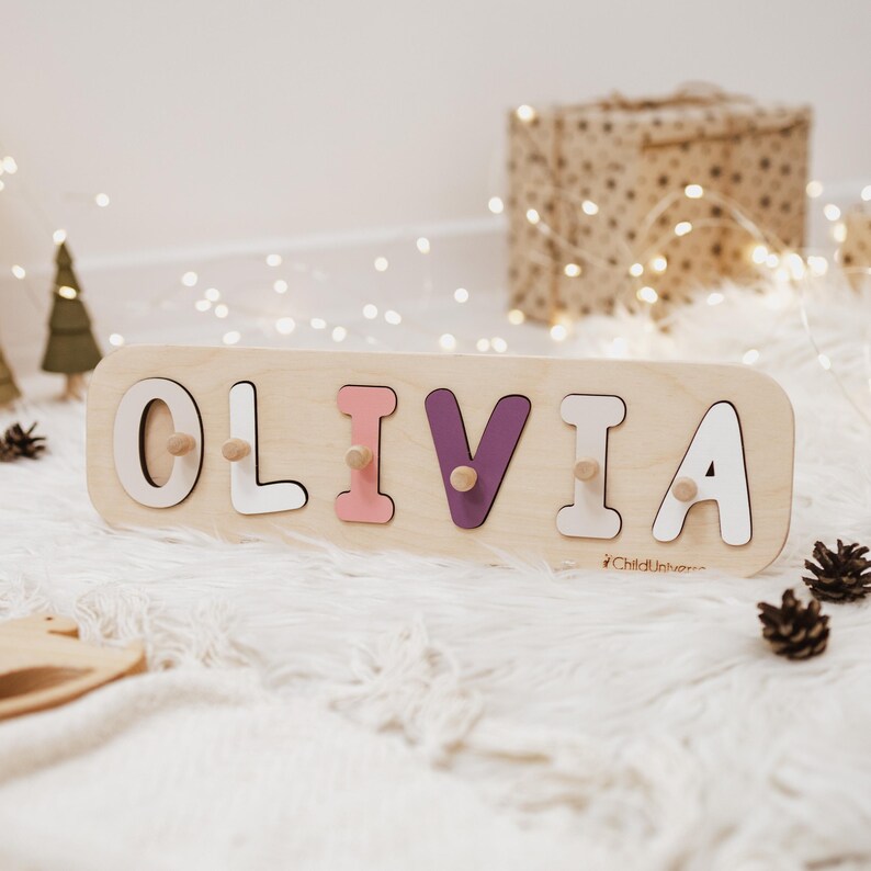 以下が含まれることがあります： 切り抜かれた文字で「OLIVIA」と書かれた明るい色の木製ネームパズルボード。文字は白、ソフトピンク、紫で色付けされており、それぞれに掴むための小さな木製のノブが付いています。ブランド名「ChildUniverse」が印刷されています。
