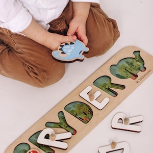 May include: Wooden dinosaur puzzle with the name "RYDER" spelled out in white letters. The puzzle pieces feature colorful dinosaur illustrations and a light wood finish. A child is holding a blue dinosaur piece.
